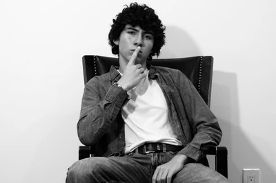 Monochrome photo of Nicholas Fortune sitting in an old embellished office chair staring into the camera with a finger over his mouth