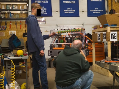 A photo of FRC Team 2013's 2026 season robot in the workshop with two people surround it